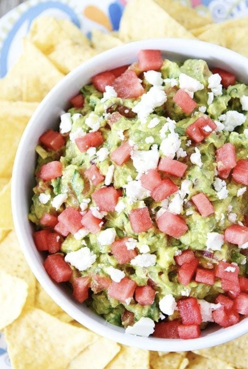 A bowl of guacamole topped with feta cheese and diced watermelon, surrounded by tortilla chips