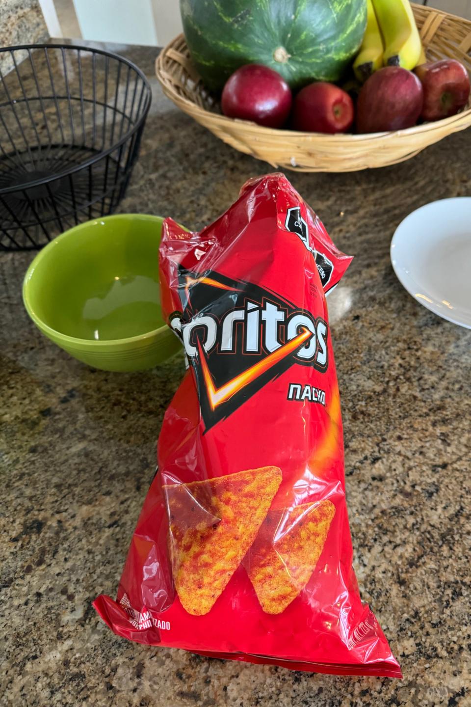 An open bag of Doritos Nacho chips on a kitchen counter with a basket containing a watermelon, bananas, and apples in the background