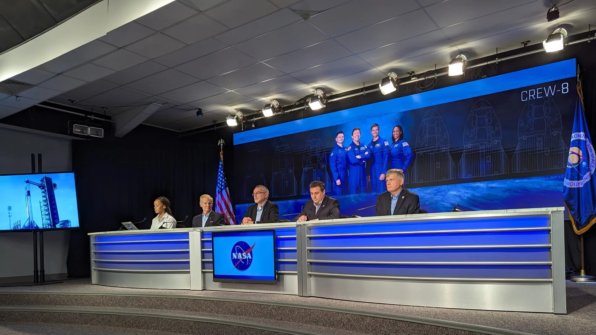  People in suits sit at a desk in front of a poster of four astronauts in flight suits and the text "crew-8". 