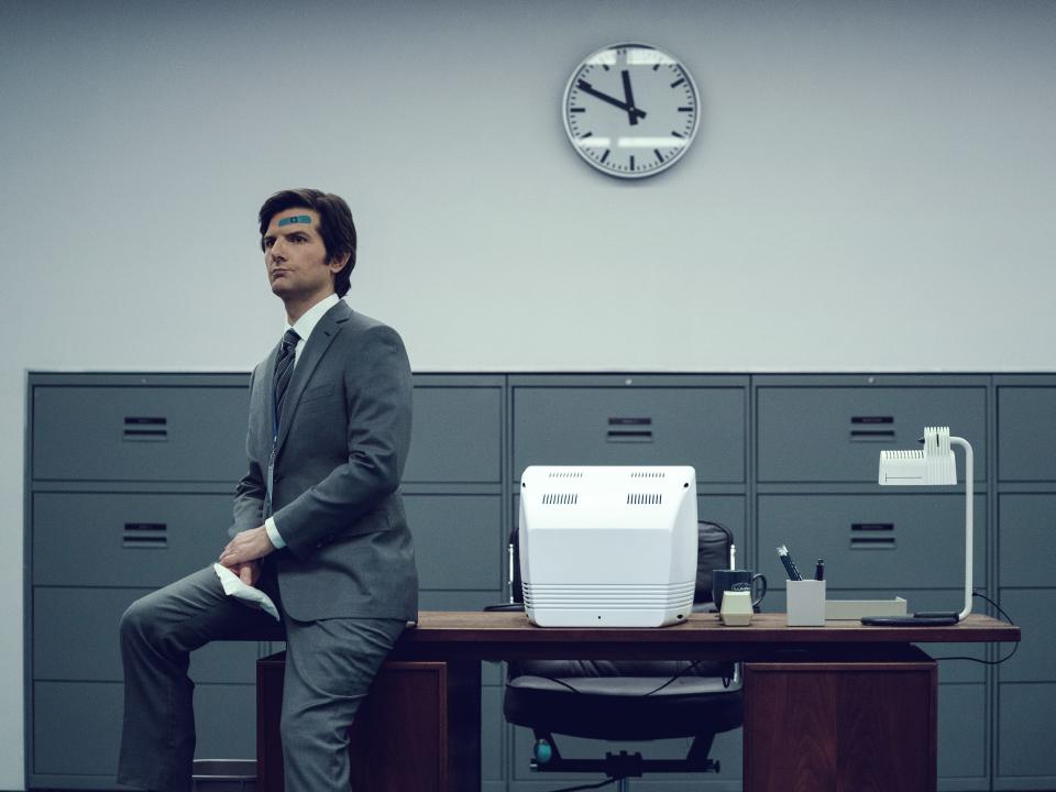 adam scott as mark in severance, sitting on a desk with an older-looking computer and a mid-height row of file cabinets in the background