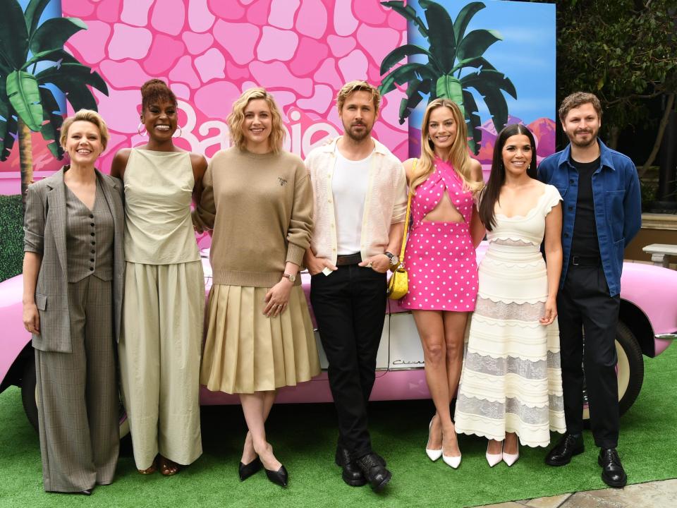 The cast of "Barbie" standing in front of a pink car while Margot Robbie wears a bright pink outfit.