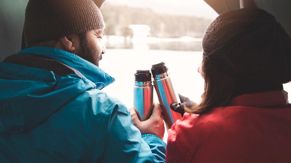 A woman and man sat in a tent cheersing their flasks