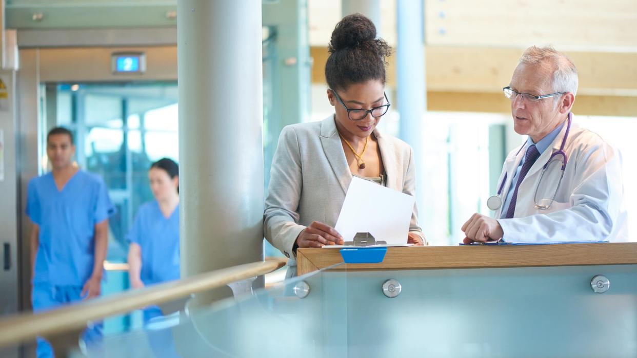 A suited woman and a male doctor wearing a lab coat stand on a stairwell of a modern hospital and discuss some case notes.