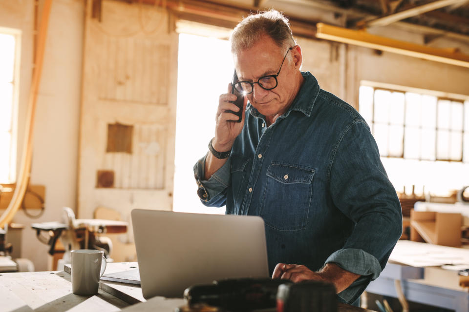 Älterer Mann, der am Handy spricht und Laptop auf dem Arbeitstisch verwendet. Reifer Tischler, der am Laptop arbeitet und in seiner Tischlerei einen Anruf entgegennimmt.