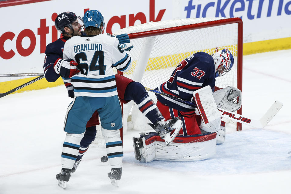 The puck trickles wide after a save by Winnipeg Jets goaltender Connor Hellebuyck (37), while Dylan DeMelo (2) defends against San Jose Sharks' Mikael Granlund (64) during the second period of an NHL hockey game Wednesday, Feb. 14, 2024, in Winnipeg, Manitoba. (John Woods/The Canadian Press via AP)