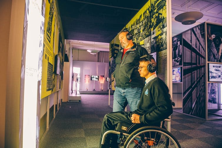 A man in a wheelchair and a man standing wear sets of headphones. Both are in a large room and are looking at a screen.