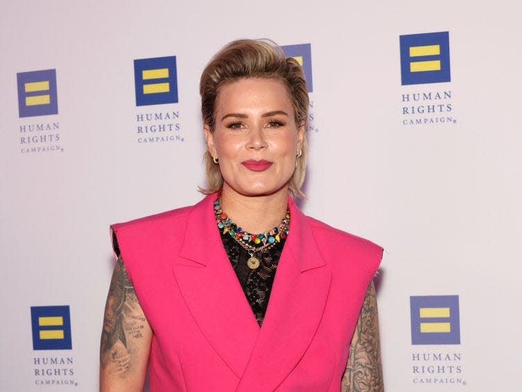 ashlyn harris on a human rights campaign red carpet, wearing a pink suit without sleeves that show her tattooed arms. her hair is worn pushed back from her forehead