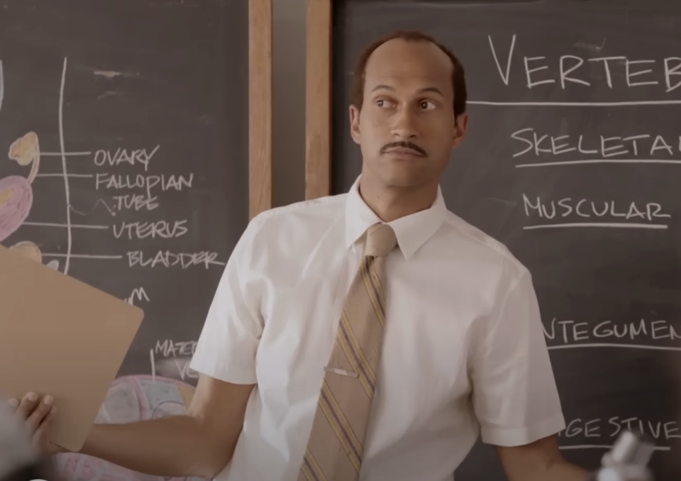 Keegan-Michael Key in a classroom scene, holding a folder with a surprised expression, a chalkboard with anatomical terms behind him