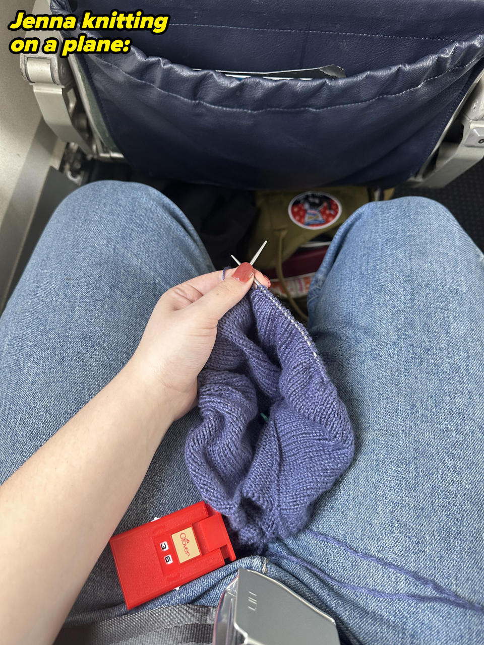 A person is knitting with purple yarn while sitting on an airplane. They have knitting needles and a red measuring tape in their lap