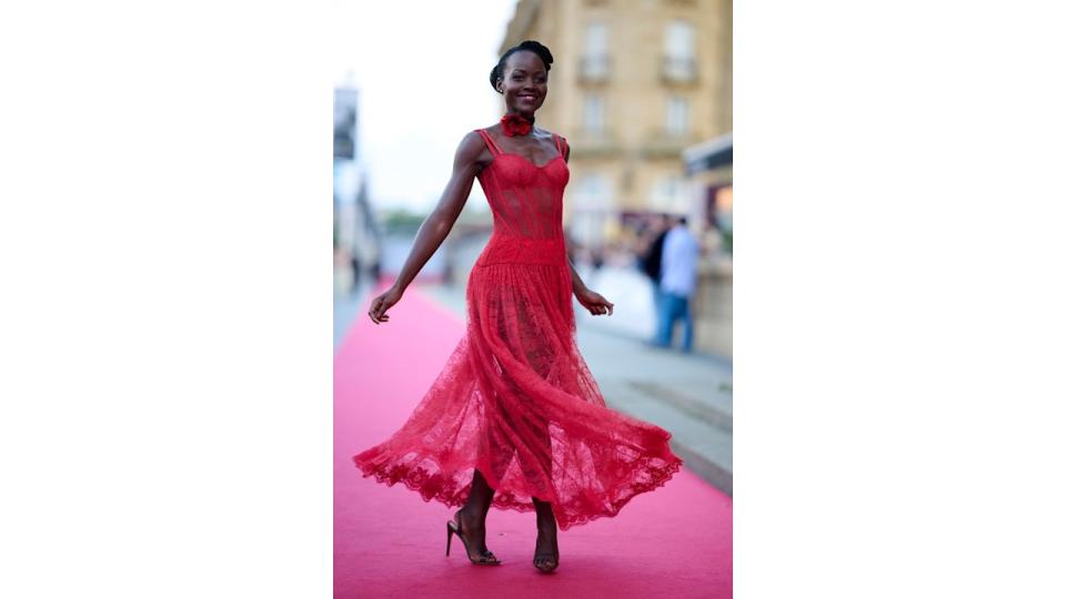 Actress Lupita Nyong'o woman twirls slightly, showcasing the movement of her red lace dress on the red carpet. The corset bodice and sheer flowing skirt catch the light. She smiles warmly, exuding confidence as the wind lightly plays with her dress.