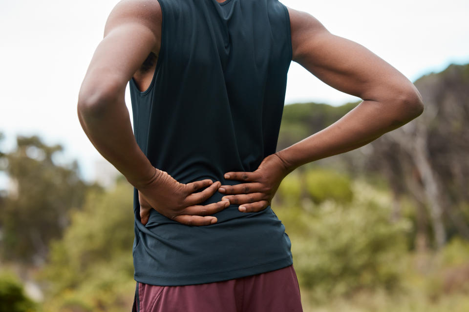 Person outdoors in athletic clothing, facing away, holding their lower back with both hands, suggesting discomfort or pain