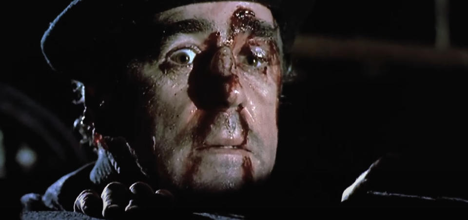 A close-up of a scared man with blood and injuries on his face, wearing a hat, from a scene in a movie or TV show. No other persons or notable text visible