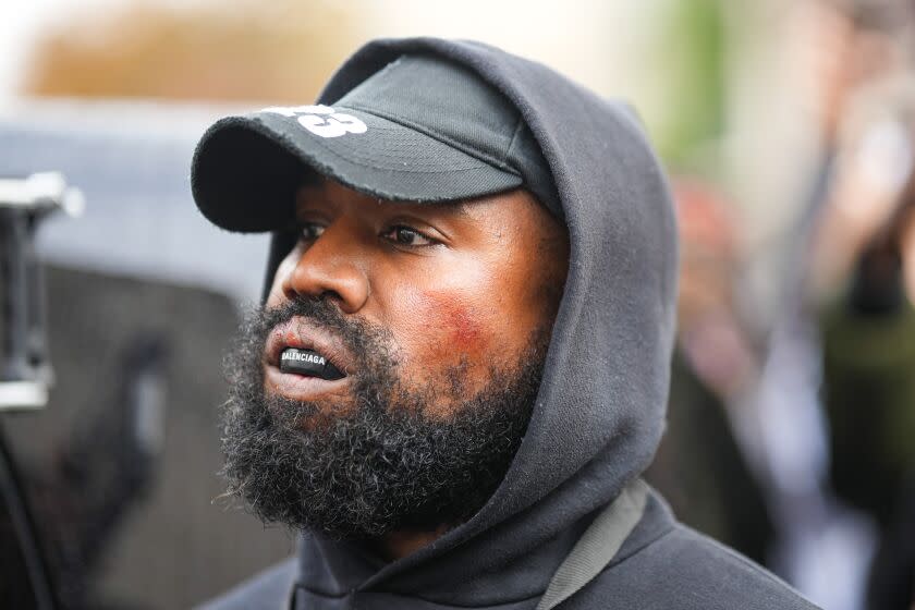 A man with a beard and mustache wears a black hoodie over a black hat with a Balenciaga boxing mouthguard