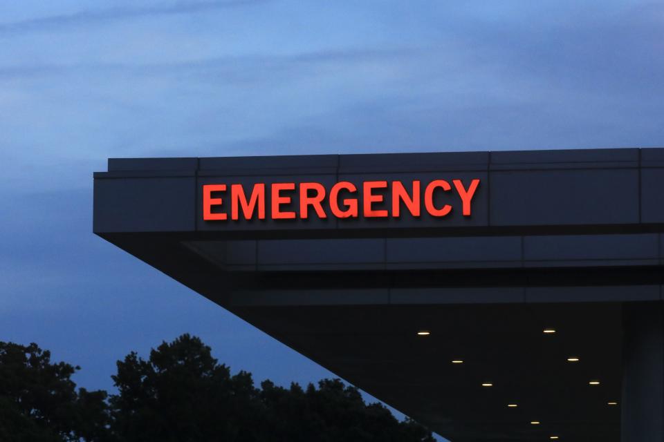 Exterior of an emergency department entrance with a red "EMERGENCY" sign illuminated against the evening sky