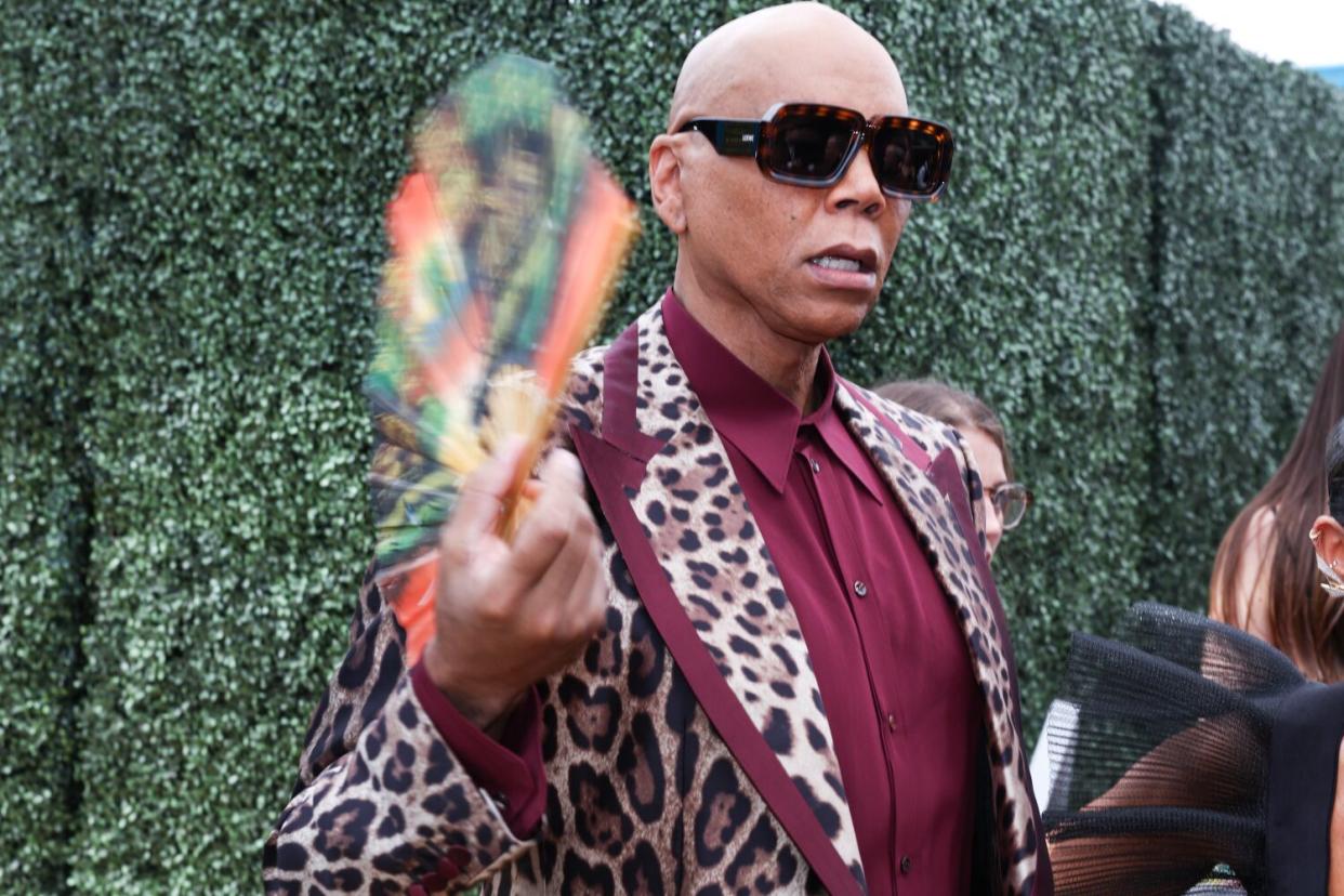 RuPaul, in a cheetah print jacket and large black sunglasses, holds up a fan