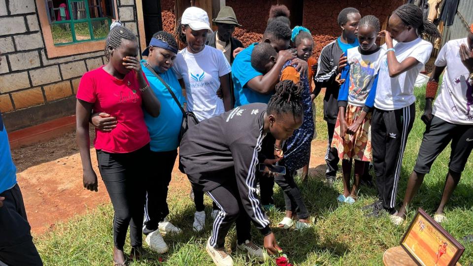 A group of people cry and hug each other as one of them places flowers on the spot next to a photo of Rebecca Cheptegei