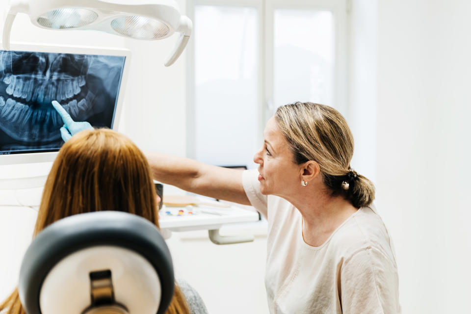 An orthodontist pointing at some teeth on an x-ray displayed on a surgery monitor while talking to a patient about a dental procedure.