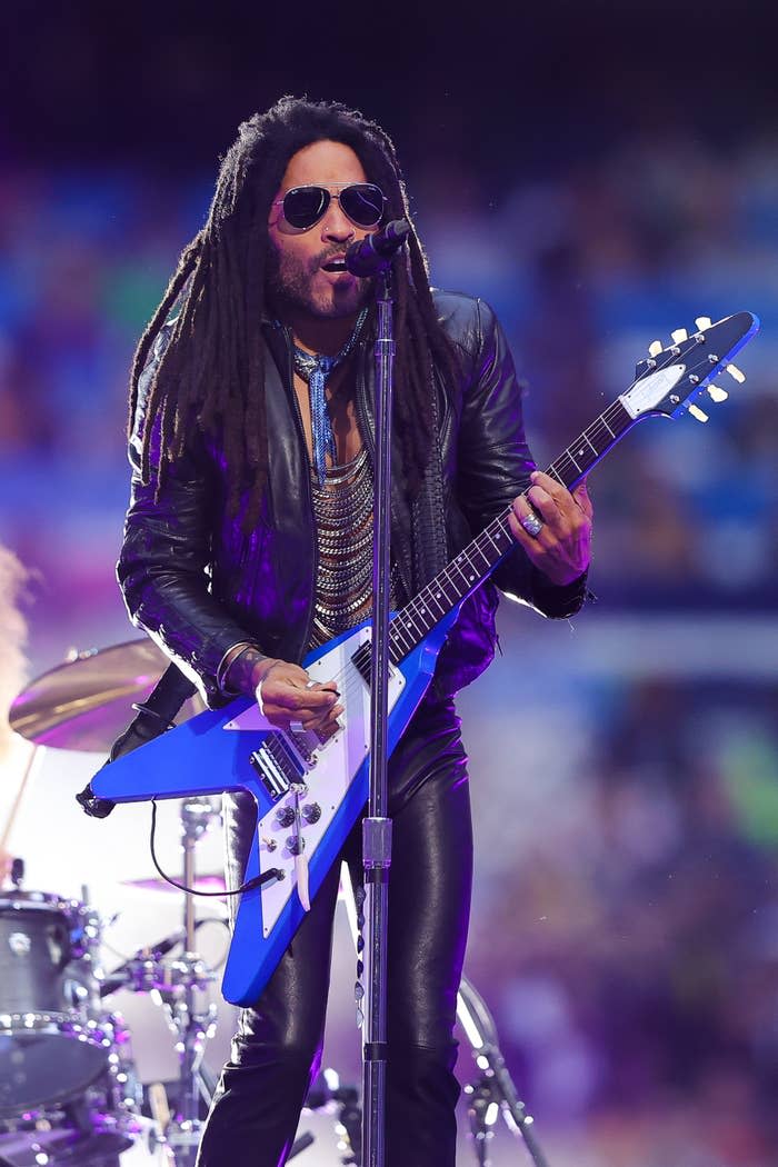 Lenny Kravitz performs on stage wearing a leather jacket, sunglasses and black pants and playing a blue electric guitar