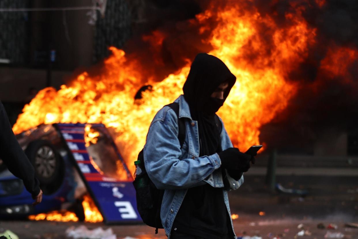 Una persona con el rostro cubierto mira su teléfono mientras arde un auto en las protestas en Buenos Aires. 