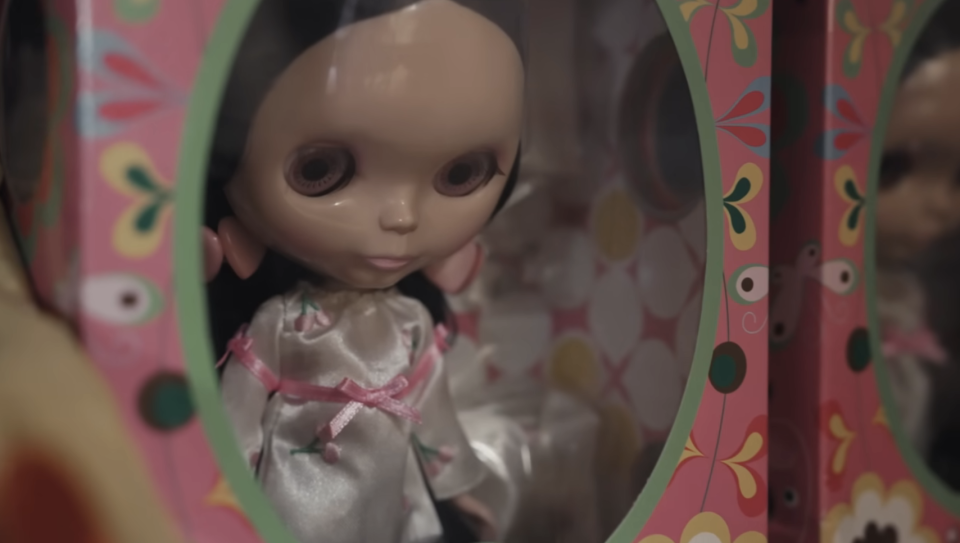 A Blythe doll with large eyes wearing a shiny dress, enclosed in a colorful, patterned box