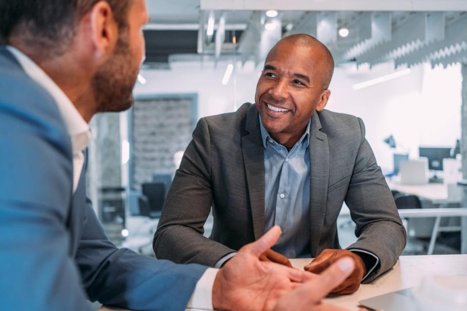 A person wearing professional attire and sitting at a table smiles while listening to another person across the table. 