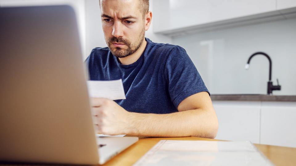 Young frowning bearded serious man sitting at dining table, holding bill and using laptop for paying it.