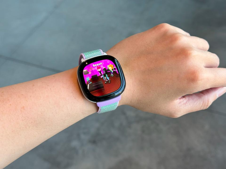 Fitbit Ace LTE, held aloft on the wrist, displays a digital character inside a pink bedroom on the screen.  Above is the word 