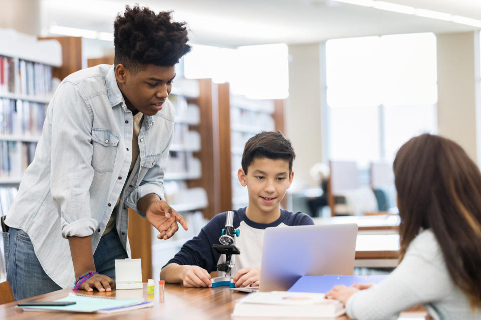 A teenage boy stands over two middle schoolers at a table in their school library.  He points to a computer monitor as he speaks.  There is a microscope on the table.