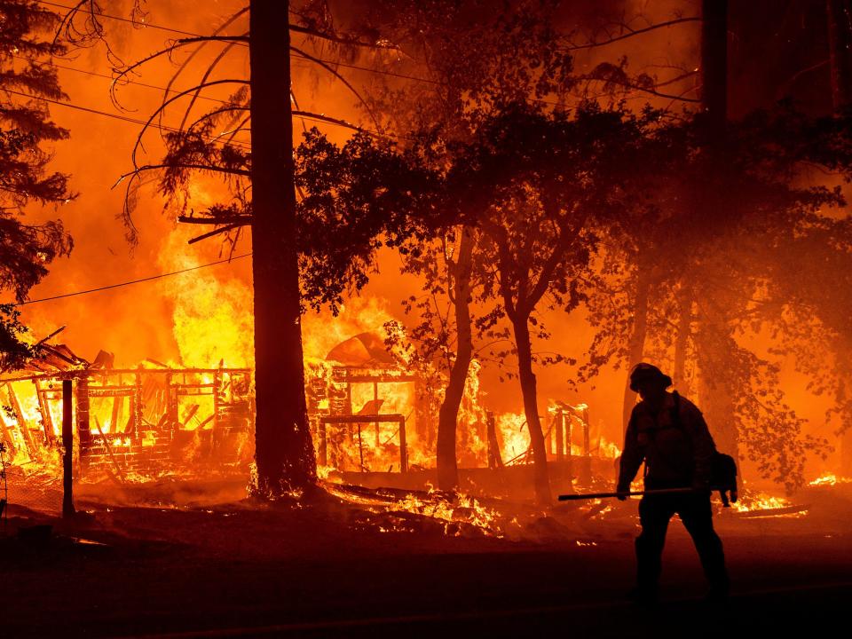 The sillouhette of a firefighter stands in front of a burning home.