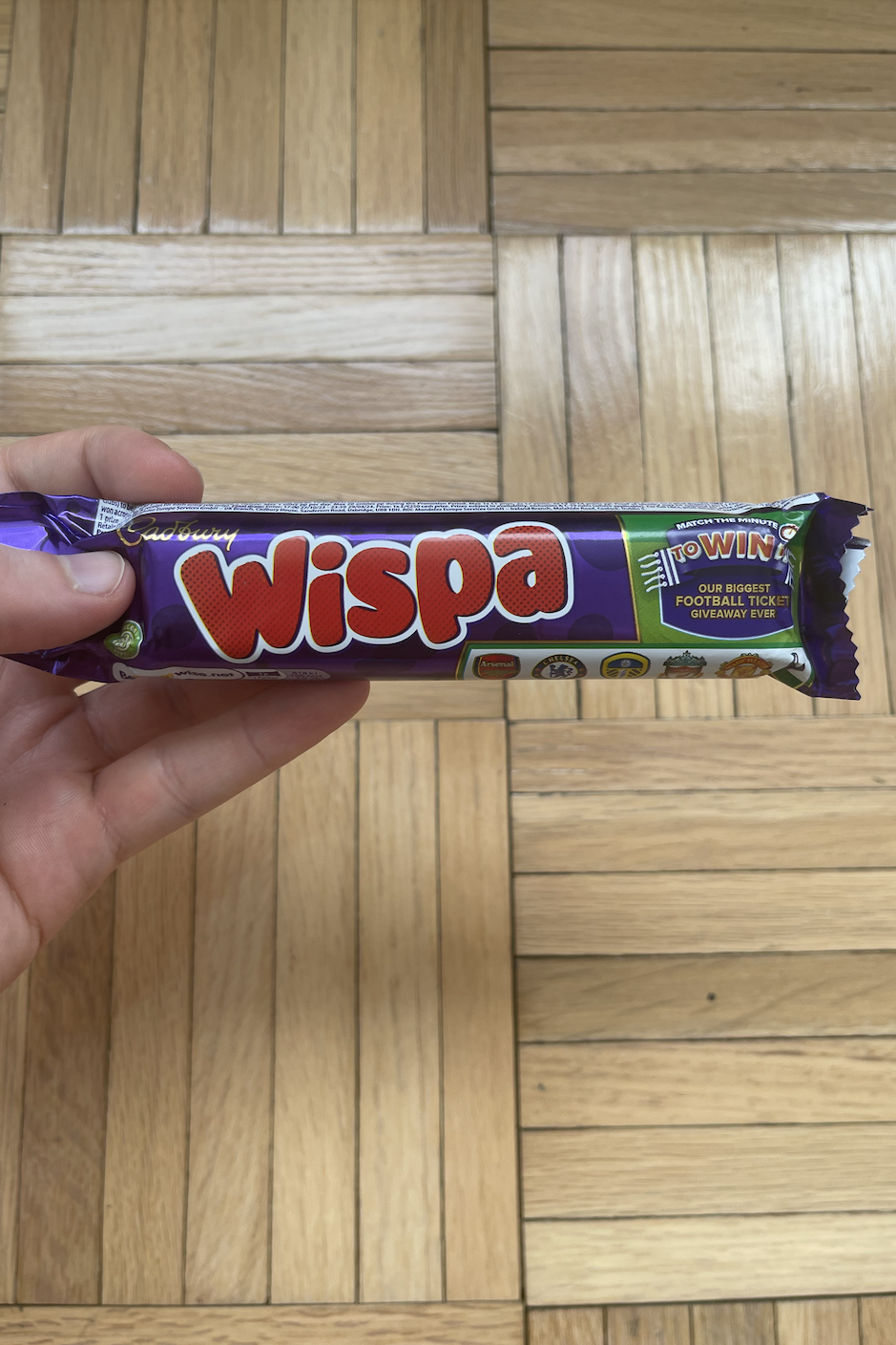 A hand holding a Wispa chocolate bar against a wooden floor background