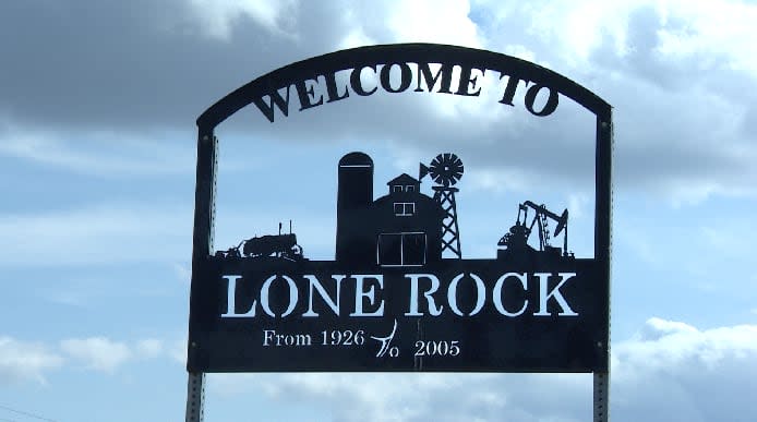 A metal signs shows a farm house, pump jack with the words "Welcome to Lone Rock: From 1926 to 2005"