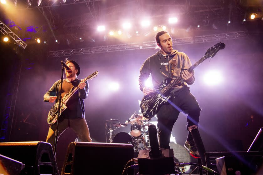 Patrick Stump holding a guitar and singing into a mic. Pete Wentz in a hoodie and pants holding a guitar. Both are on a stage