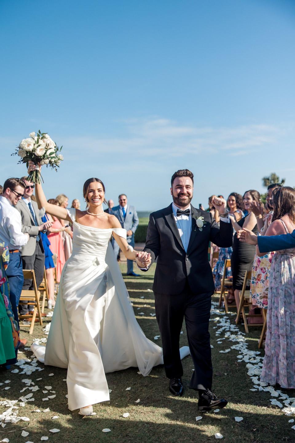 A bride and groom raise their hands in excitement as they leave their wedding ceremony.