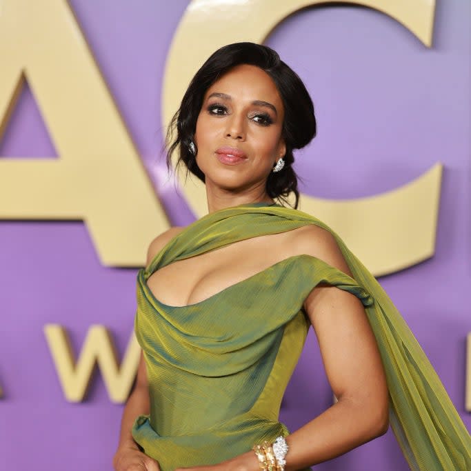 Person posing on the red carpet in an off-shoulder, draped green gown with a statement bracelet