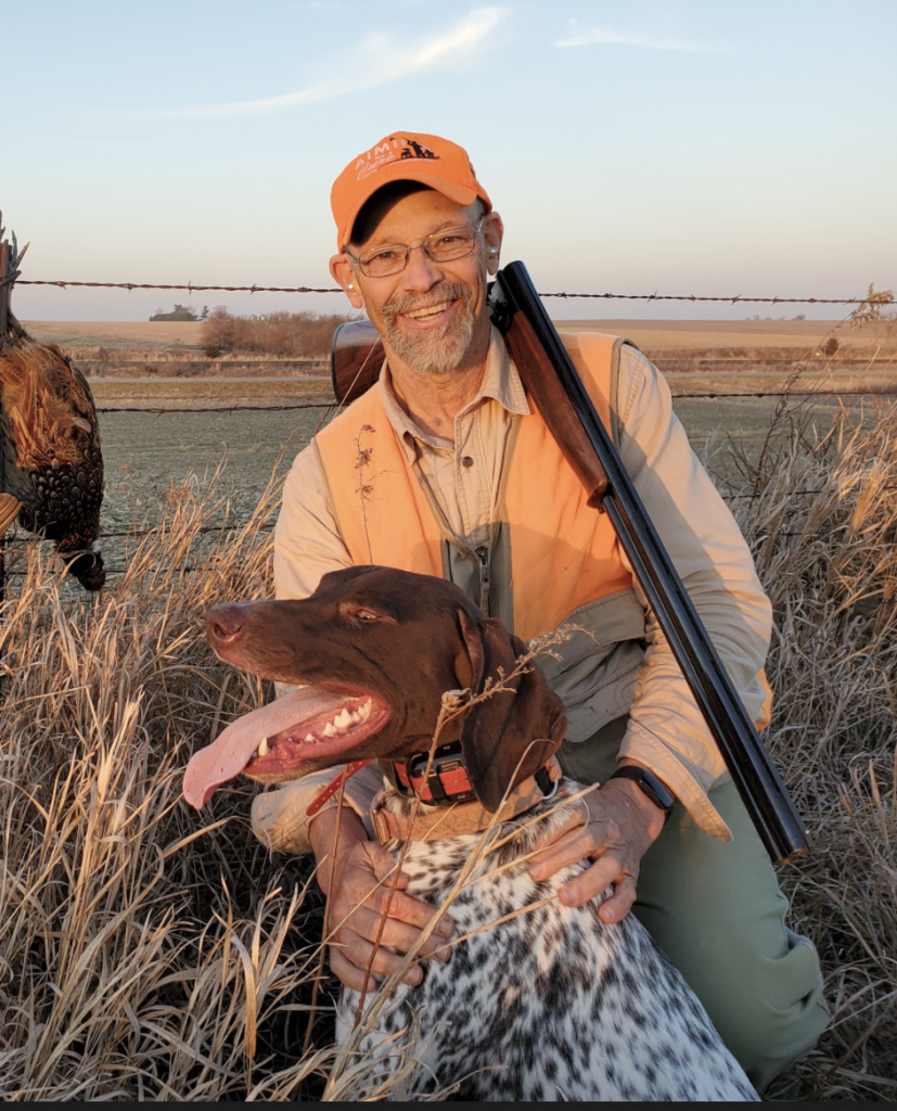 A hunter dressed in tan and orange hunting close knees beside a german shorthaired pointer