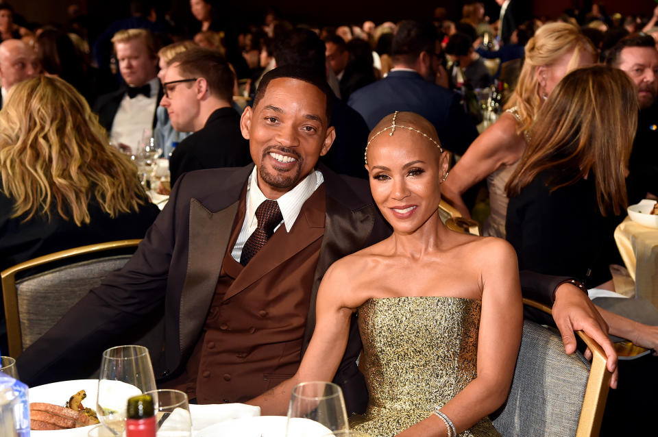 Will and Jada seated at an event, smiling; he's in a suit and tie, she's in a glittering sleeveless dress