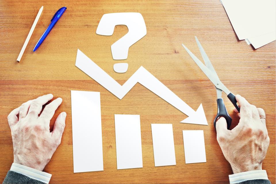 A man holds a pair of scissors in one hand, in front of a paper cutout design of a question mark, bar chart, and declining arrow.