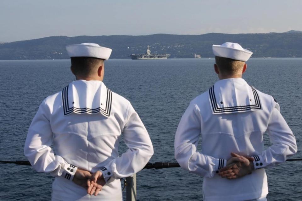 Navy sailors in white uniforms stand with their hands behind their back as they observe as military aircraft carrier anchoring in the background