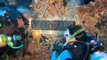 In this Sunday, May 15, 2022, photo provided by the Florida Keys News Bureau, Karen Berrios, right, watches Jon Hazelbaker install a commemorative plaque on the Spiegel Grove, a decommissioned 510-foot-long Naval Landing Ship Dock that was sunk in 2002 to become an artificial reef in the Florida Keys National Marine Sanctuary off Key Largo, Fla. Installation of the plaque was one of several activities that are being staged to mark the 20th anniversary of the sinking. Barrios' father and uncle served on the Spiegel Grove. (Frazier Nivens/Florida Keys News Bureau via AP)