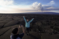 Kelly Ann Kobayashi raises her hands as she poses for a picture for Chad Saito, left, while standing on hardened lava rock from a previous eruption as the Mauna Loa volcano erupts, behind, Wednesday, Nov. 30, 2022, near Hilo, Hawaii. The two were visiting from Honolulu. (AP Photo/Gregory Bull)