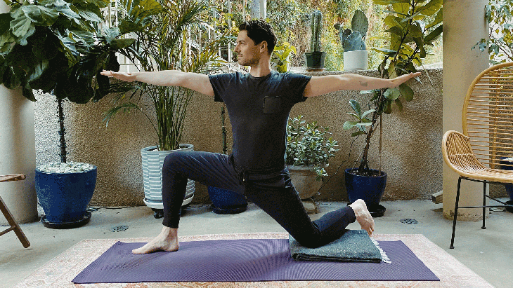 Man kneeling on a yoga mat with one knee down and the other in a lunge with his arms reaching straight out from his shoulders in Warrior 2 Pose variation.