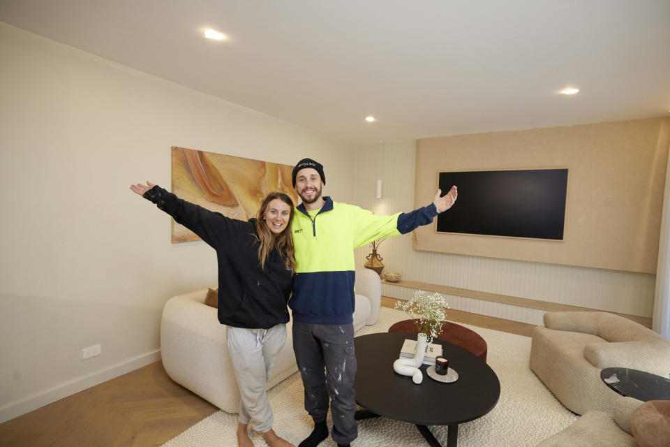 Kristy and Brett pose smiling with their arms held out in their living room.