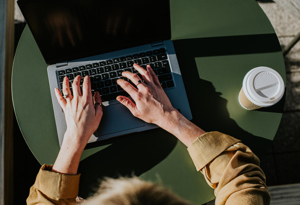 top-down view of two hands typing on a laptop. the laptop is on what seems like a café's table outside, with a disposable coffee cup beside it