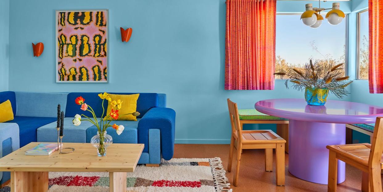 colorful living room with light blue walls and a dark blue sofa and a purple pedestal table and wood chairs and burnt orange curtains on half size window and a colorful rug under the cocktail table