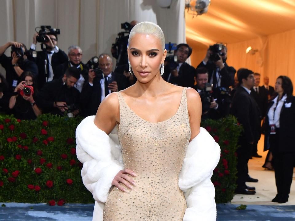 Kim in a nude fitted gown with a scoop neckline and thin straps with chest padding partially seen through the crystal-embellished fabric. She has a white fur stole worn around her arms.