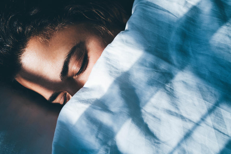 A woman's face peeking out from behind the covers of her bed, eyes closed, sunlight dancing on her skin