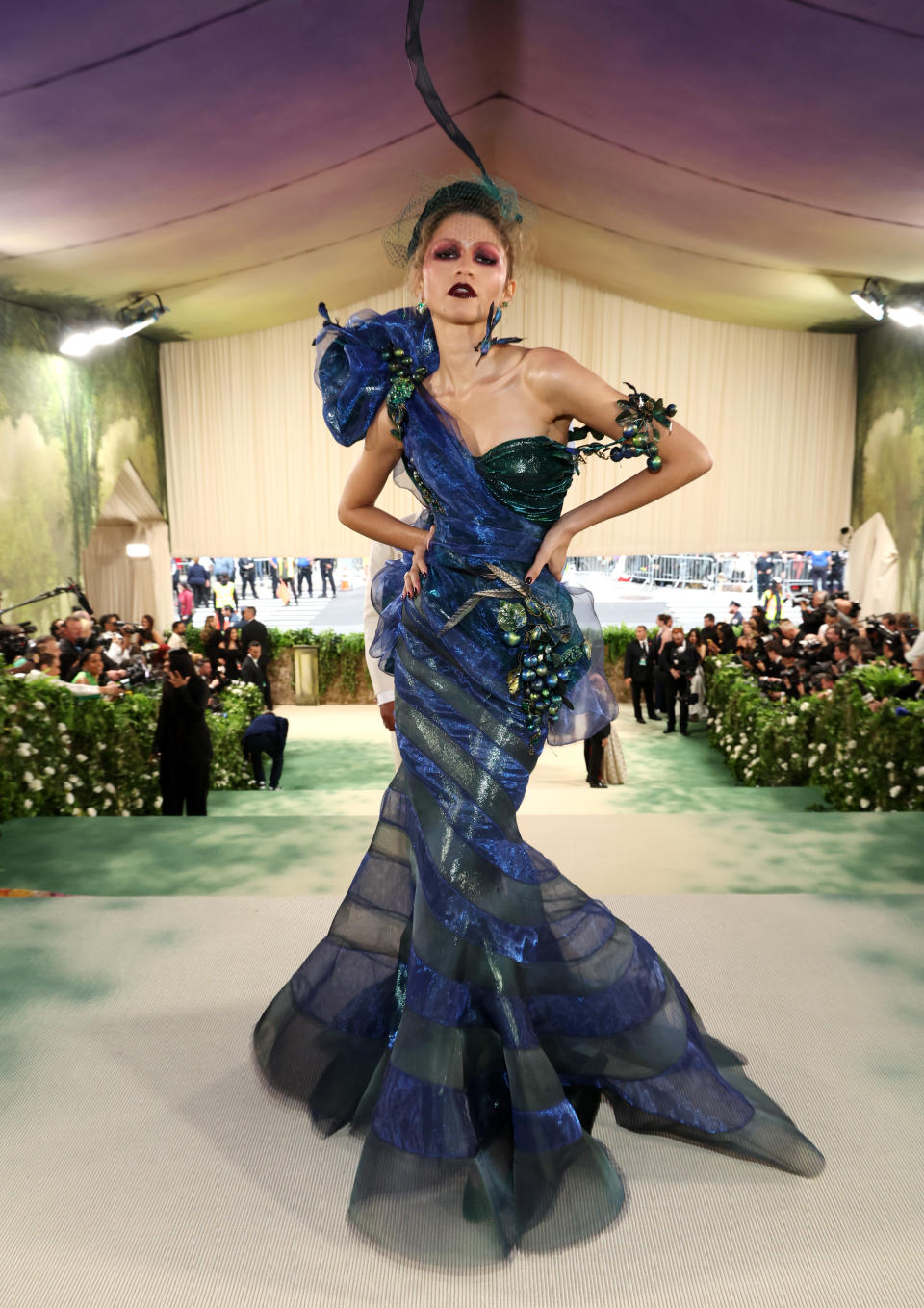Zendaya in elaborate blue-toned gown with ruffled details and headpiece, posing on a staircase at an event