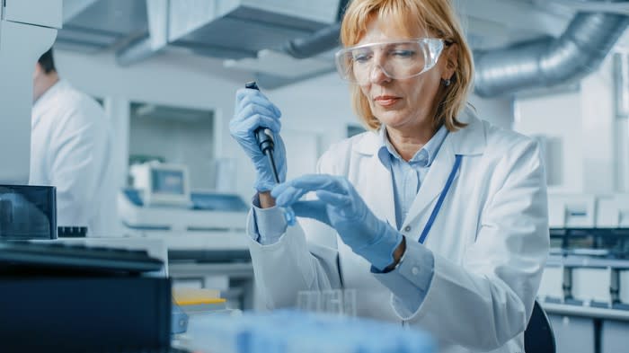 A woman in a lab coat and safety glasses uses a pipette in a lab.