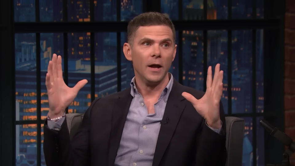 Mikey Day holds his hands up while in conversation on the set of Late Night with Seth Meyers.