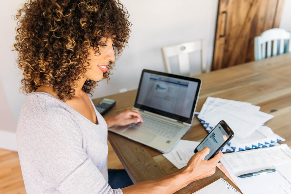 A woman sits at her dining room table with laptop and financial reports doing her monthly budget. She is smiling at the ease of use as she works on her smart phone banking app to do monthly finances, pay taxes and save money for the future.